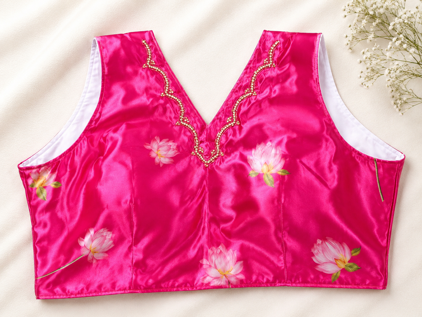 A bright pink satin sleeveless blouse top with embroidered pink lotus flowers and gold beaded scalloped trim along the V-neckline, laid flat on a cream fabric background.