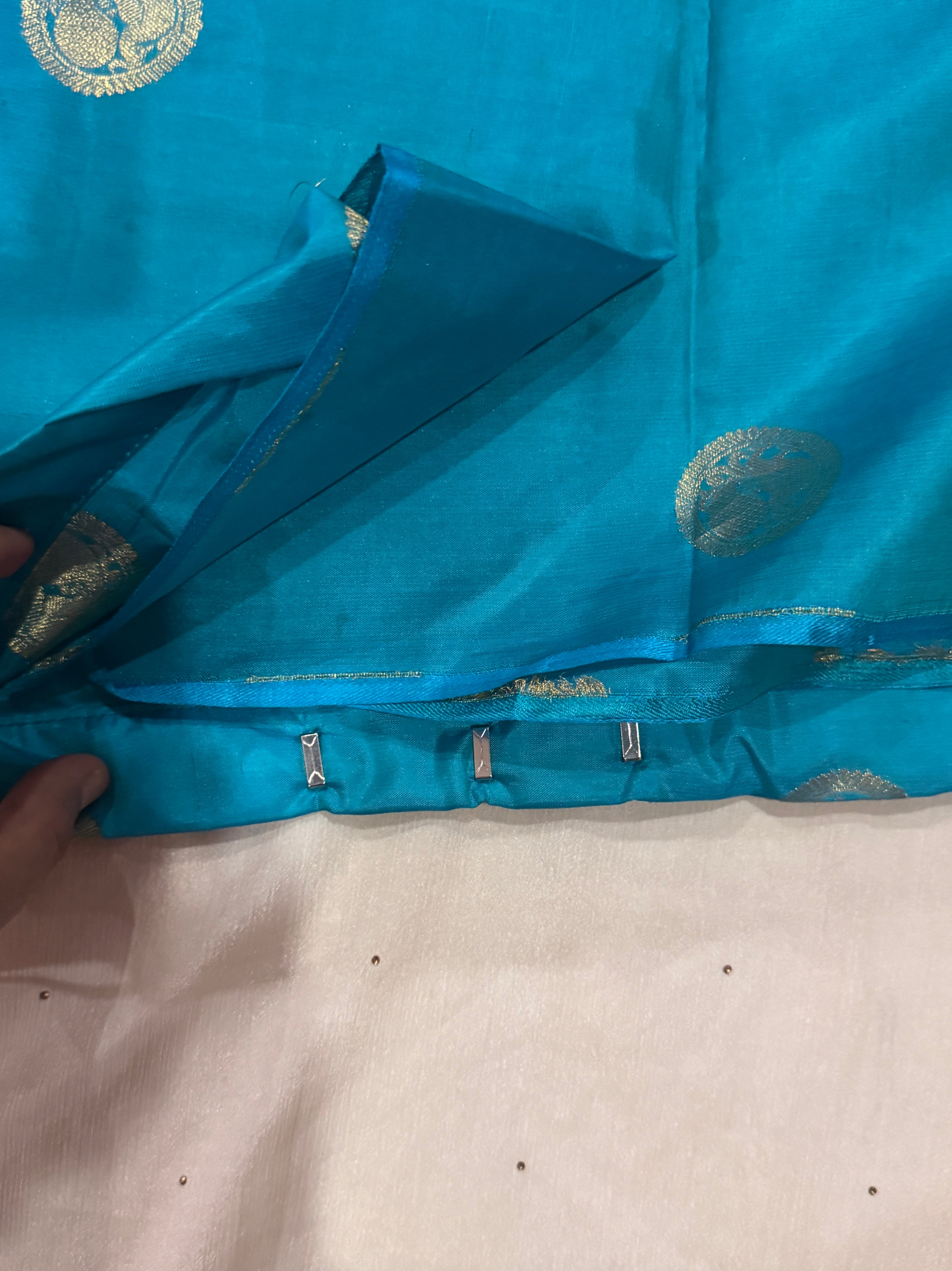 A close-up of a turquoise blue fabric garment, likely a saree or sari, featuring metallic gold zari motifs and a hand lifting the fabric to show the inner lining and edge construction.