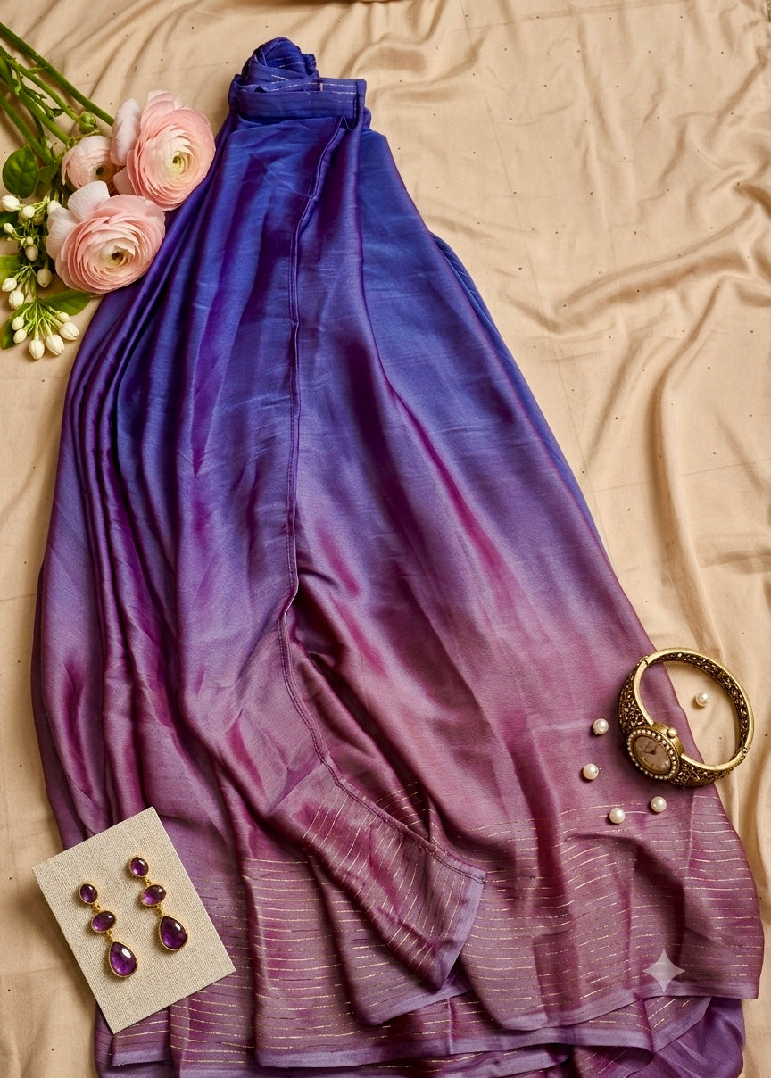 A draped scarf with a purple and blue ombré pattern, styled in a flat-lay arrangement with pink flowers, a gold bracelet, and purple earrings on a beige fabric background.