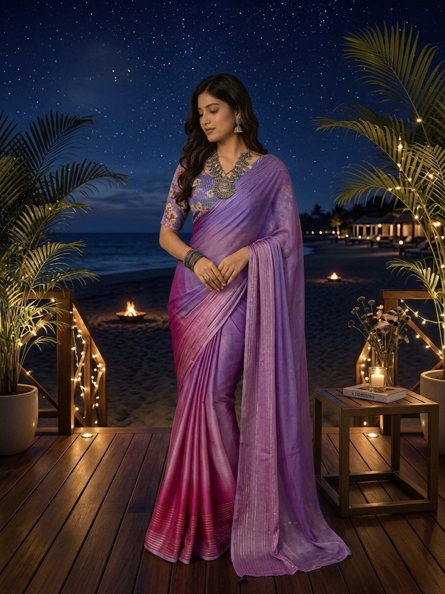A woman wearing a purple and pink gradient saree with a heavily embroidered neckline stands on a wooden deck at night, with a beach, ocean, and starry sky in the background.